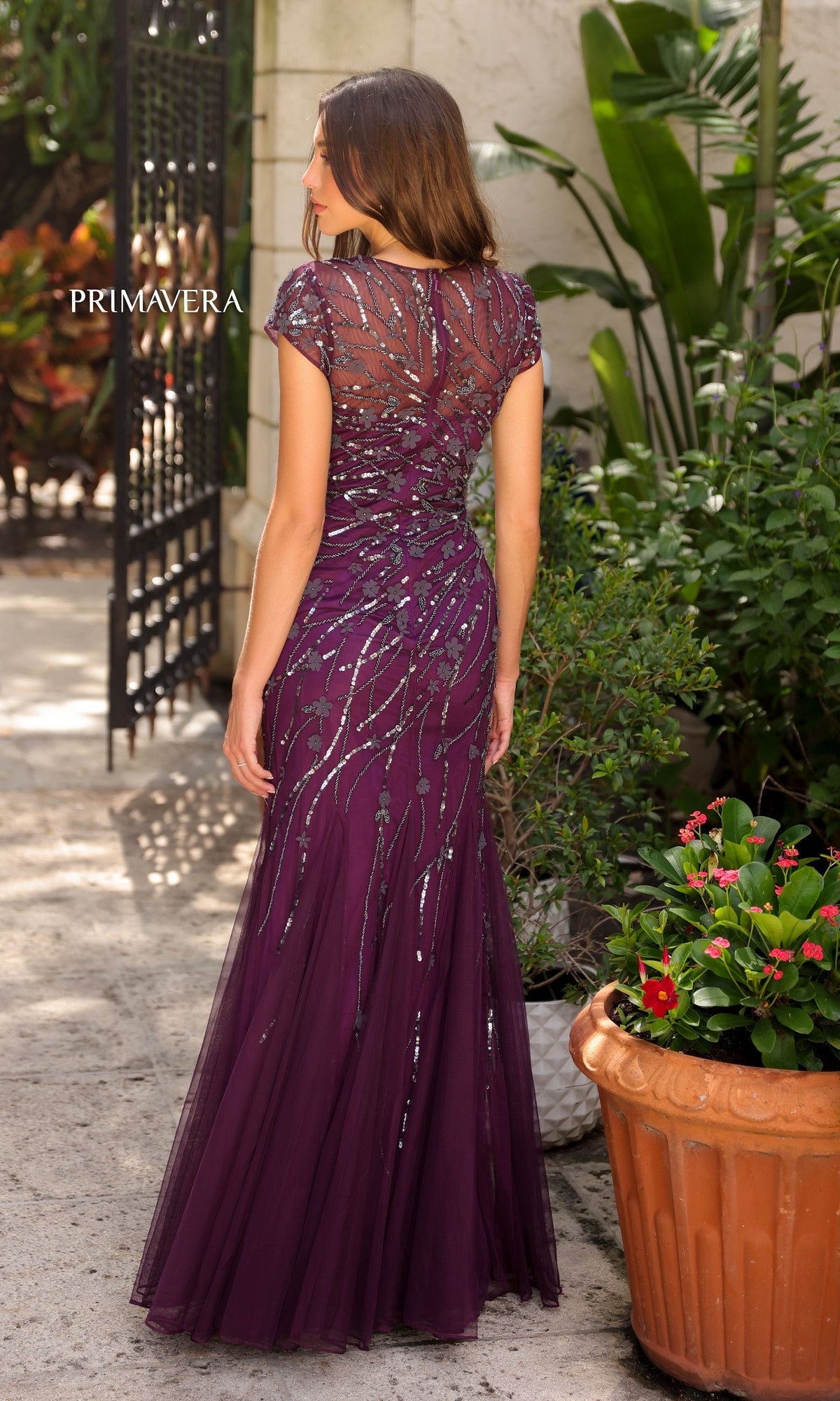 A woman stands outdoors among lush plants and flowers wearing the Long Prom Dress 13114 by Primavera—a floor-length purple gown with cap sleeves and sparkling sequin details.